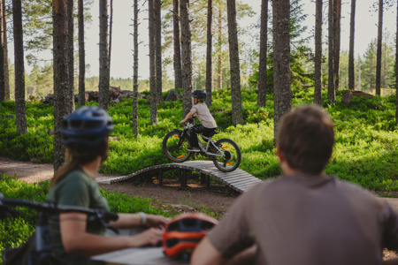 Ett barn cyklar på en teknikbana i skogen medan två vuxna tittar på från sidan.